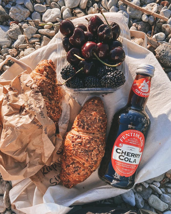 A sunny outdoor picnic scene featuring seeded croissants, fresh fruits, and a bottle of cherry cola on a rocky surface.